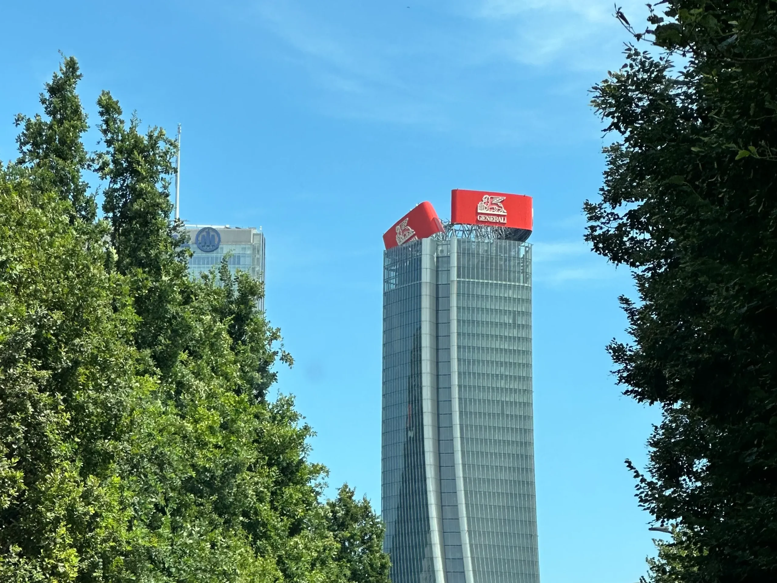 Un alto e moderno grattacielo dal design curvilineo con insegne Generali di colore rosso vivo in cima, parzialmente incorniciato da alberi verdi sotto un cielo azzurro e limpido. Un altro edificio è visibile sullo sfondo.