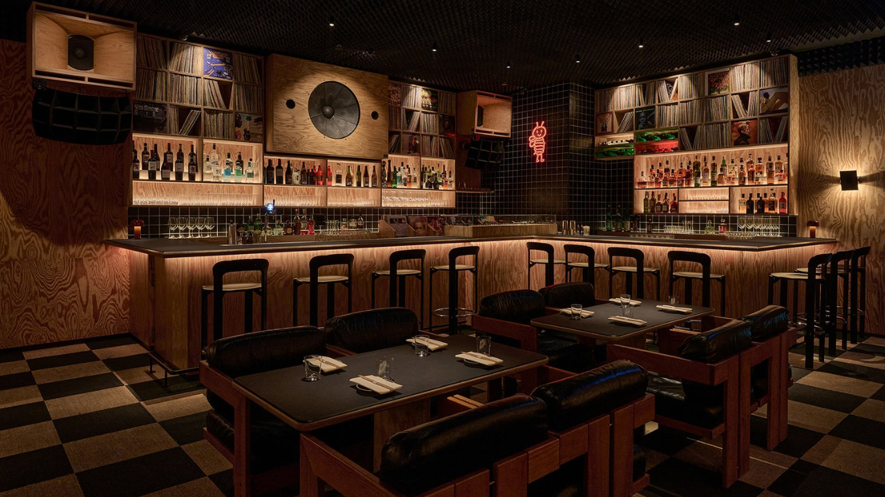 Un bar di Dubai poco illuminato con pareti in legno, scaffali di bottiglie di liquore e dischi dietro il bancone, sedie nere imbottite, tavoli quadrati con tovaglioli e un pavimento a scacchi neri e marroni.