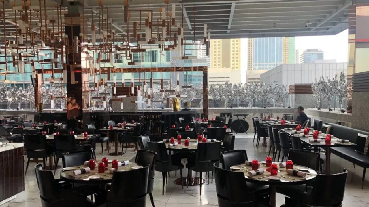Un ristorante moderno e spazioso a Dubai, con sedie nere, tavoli rotondi apparecchiati con tovaglioli rossi e soffitto decorato in oro. Le grandi finestre mostrano gli alti edifici della città all'esterno. Pochi commensali sono seduti all'interno.