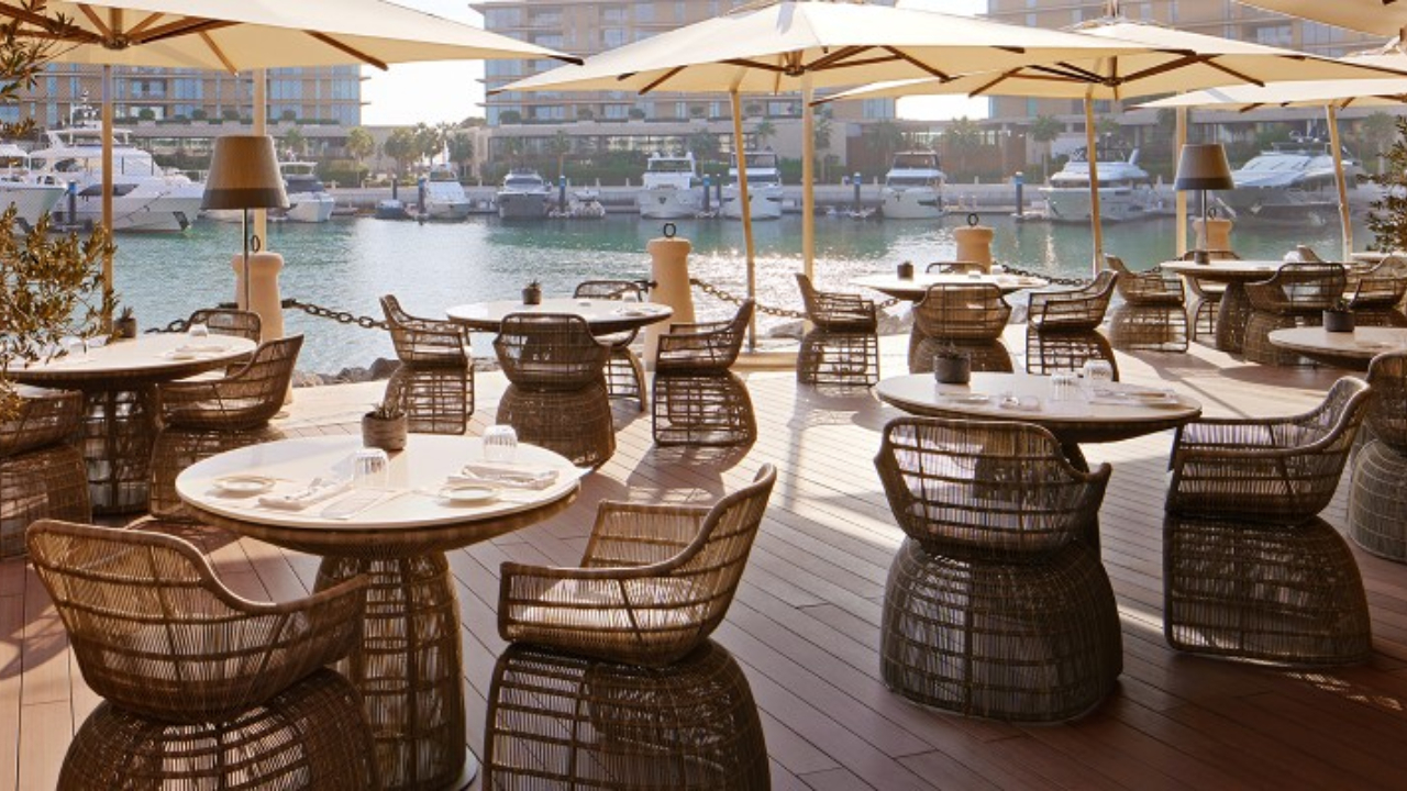 Patio del ristorante all'aperto con tavoli rotondi e sedie in vimini sotto grandi ombrelloni, con vista su un porto turistico di Dubai pieno di yacht ed edifici moderni sullo sfondo in una giornata di sole.