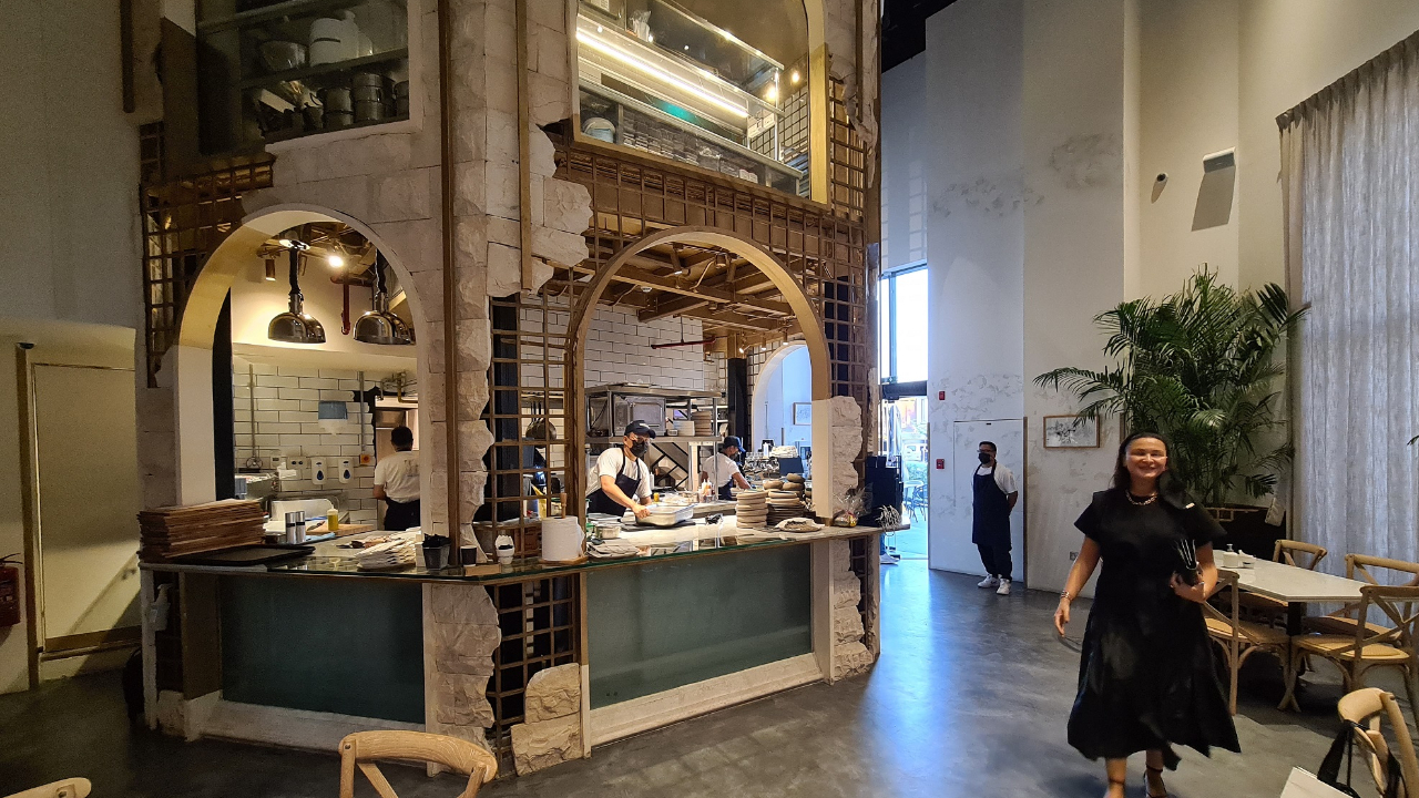 Interno di un moderno ristorante di Dubai con soffitti alti e aperture ad arco che mostrano la cucina. Il personale lavora dietro il bancone, mentre una donna in abito nero entra. Sedie e tavoli in legno riempiono l'elegante zona pranzo.
