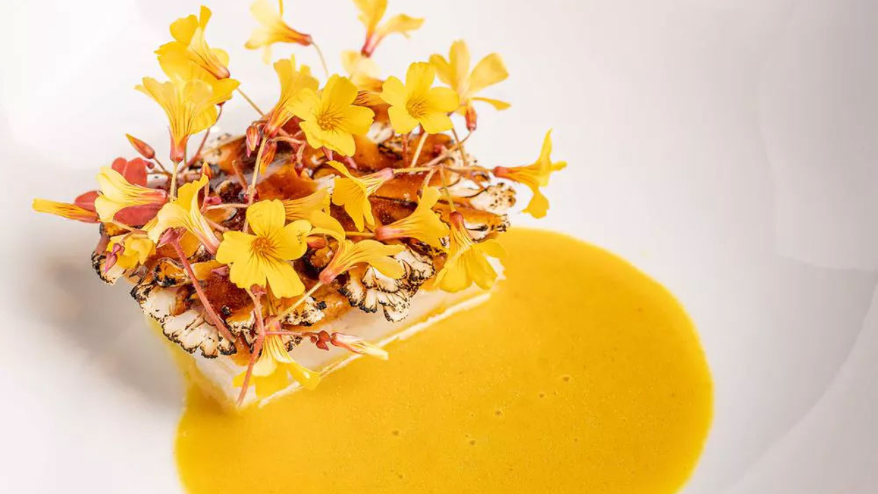 Un piatto gourmet ispirato a Dubai, con cavolfiore grigliato sormontato da fiori commestibili di colore giallo vibrante e servito con una salsa gialla cremosa su un piatto bianco.