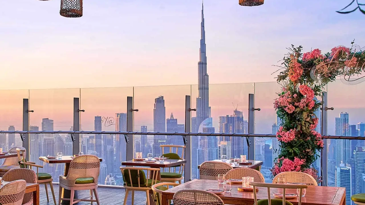 Ristorante sul tetto con tavoli e sedie in legno, con vista sullo skyline della città al tramonto. Il Burj Khalifa è visibile sullo sfondo e fiori vivaci decorano la ringhiera della terrazza.