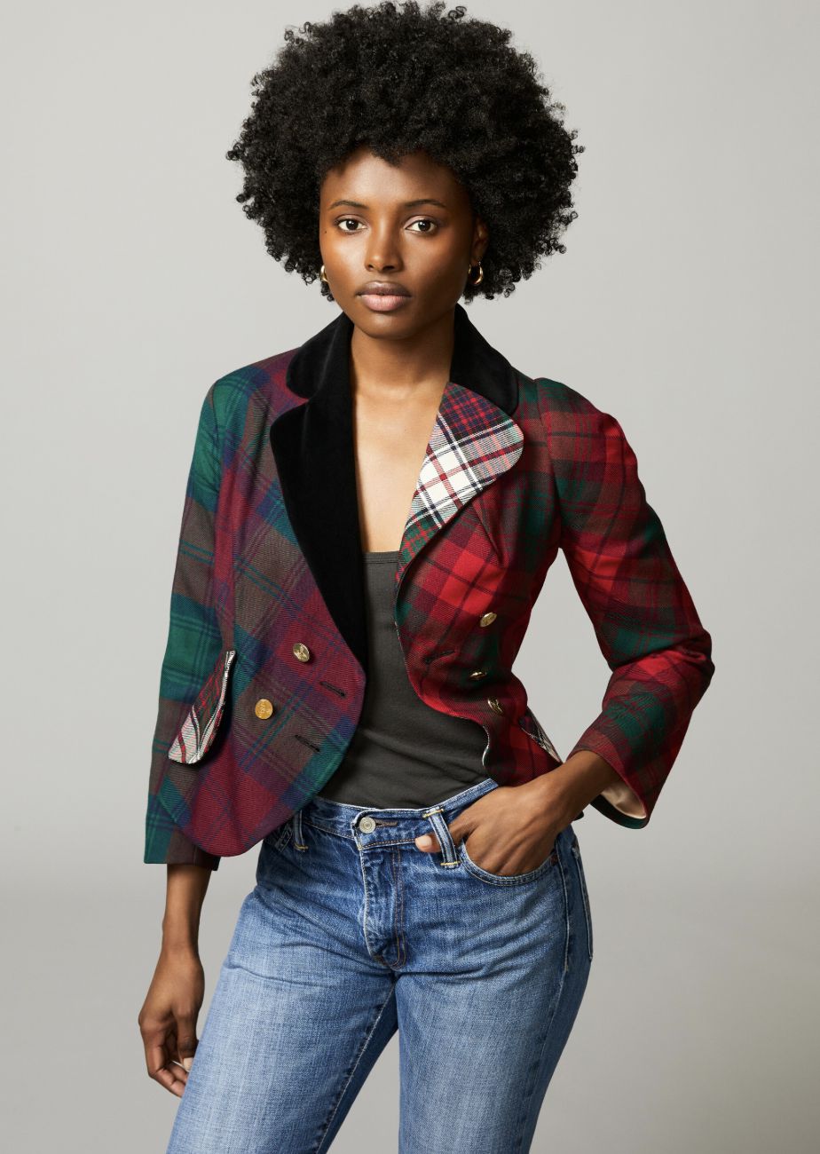 Una donna con un'acconciatura afro naturale indossa un blazer a quadri patchwork colorato su un top scuro e jeans blu, accessoriato con una borsa chic Jane Birkin, su uno sfondo grigio semplice.