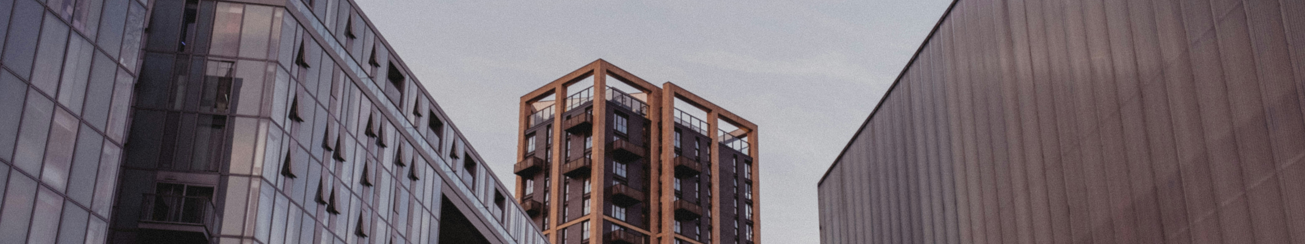 Tra due grandi edifici in vetro e metallo, sotto un cielo nuvoloso, sorge un moderno e spigoloso edificio di appartamenti con struttura in legno.