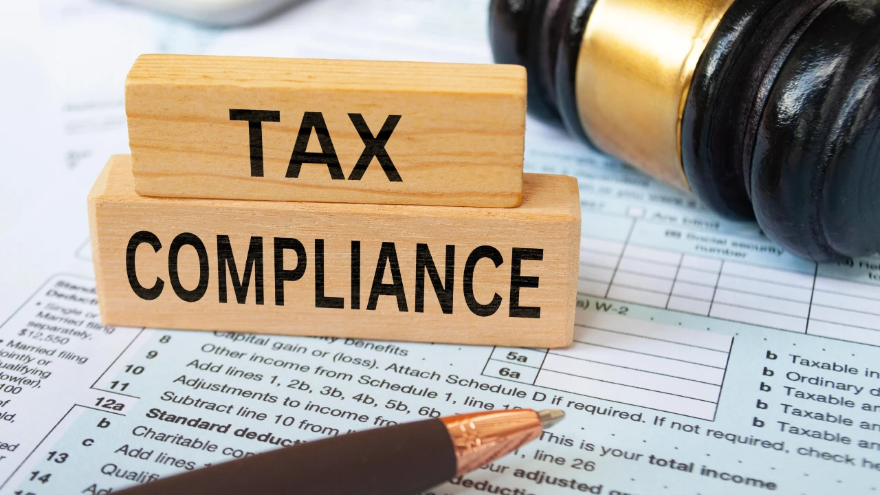 Un blocco di legno con la scritta "tax compliance" si trova sui moduli fiscali, accanto a una penna e a un martelletto da giudice, evidenziando il ruolo dell'adempimento collaborativo tra fisco e imprese