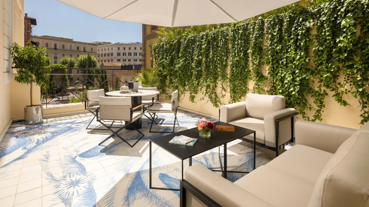Una terrazza illuminata dal sole con divani color crema, un tavolino nero, un tavolo da pranzo rotondo con sedie, piante in vaso e una parete coperta di viti verdi sotto un grande ombrellone bianco. Sullo sfondo si intravedono degli edifici.