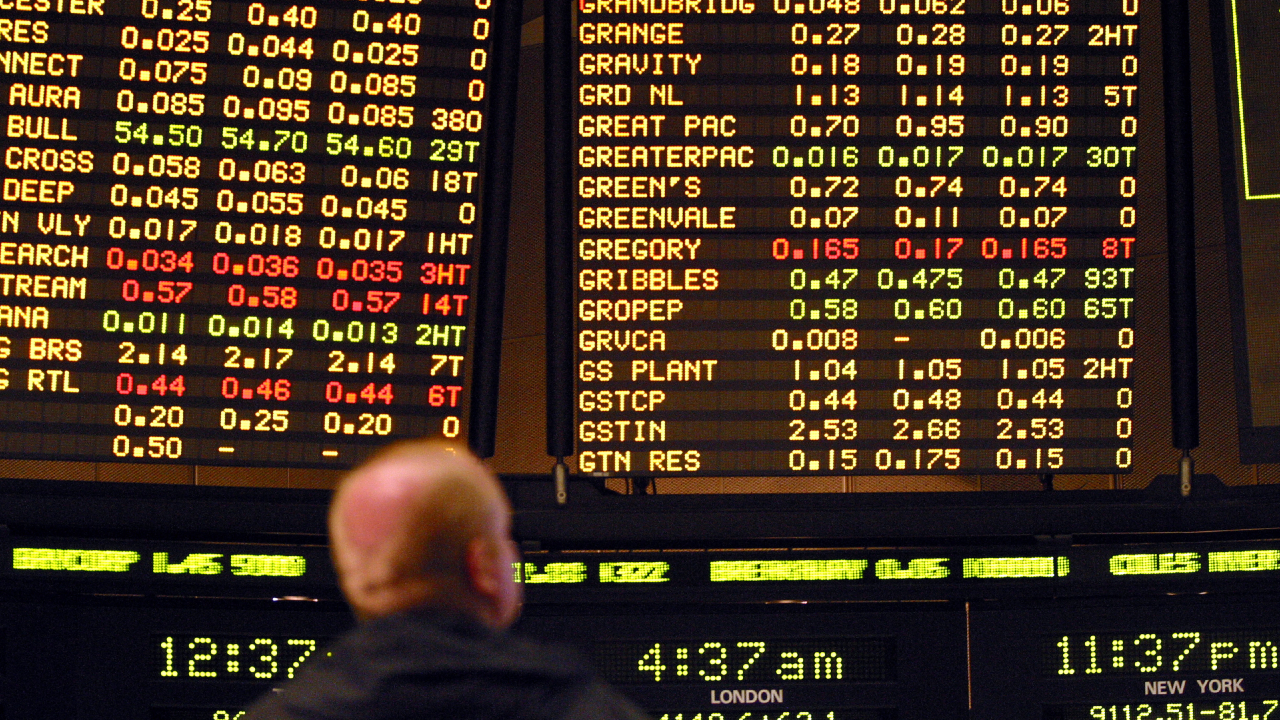 Una persona guarda un grande tabellone elettronico che mostra file di informazioni sulle azioni o sulle scommesse, con nomi di società, numeri e valori in rosso, giallo e bianco su sfondo nero.