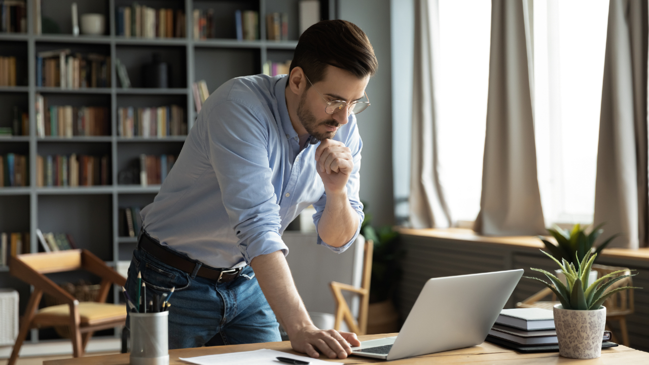 Un uomo con occhiali e camicia blu si appoggia a una scrivania e guarda pensieroso un computer portatile. Sullo sfondo ci sono librerie e grandi finestre, mentre sulla scrivania ci sono piante e materiale da ufficio.