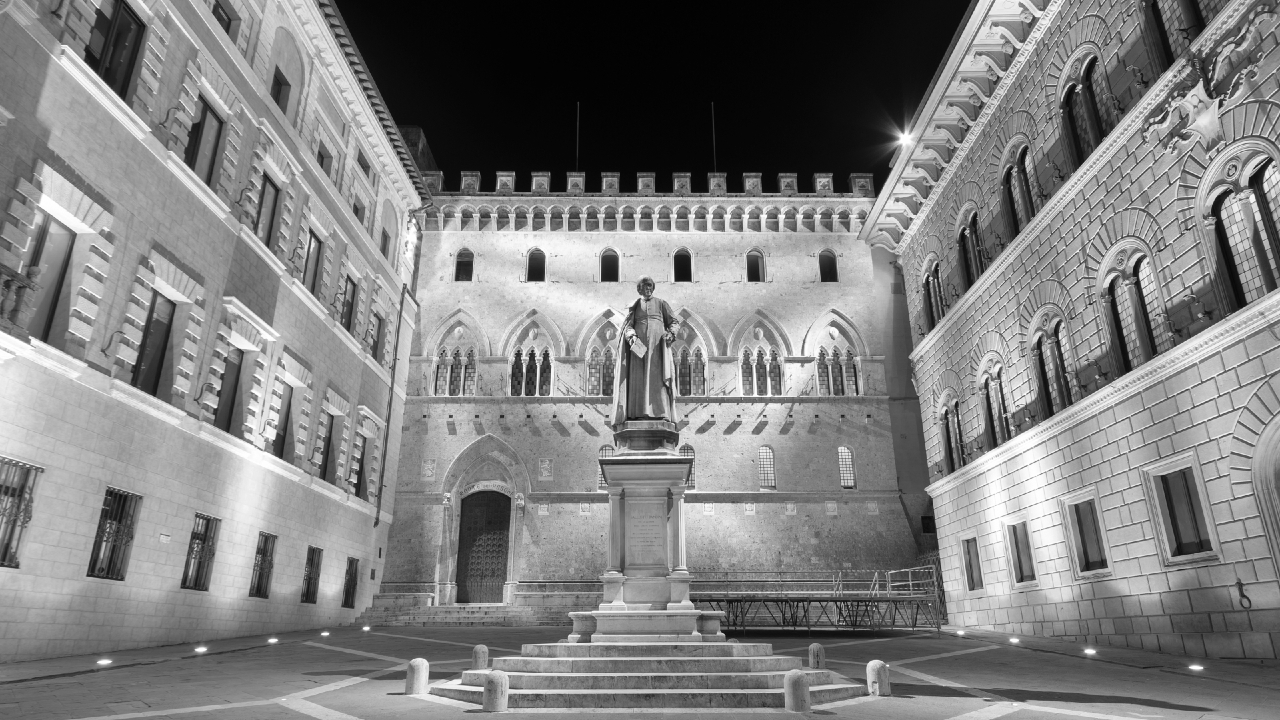 Una veduta notturna di una piazza storica con una statua al centro, circondata da edifici in pietra ornati e illuminati con finestre ad arco e facciate dettagliate. L'immagine è in bianco e nero.