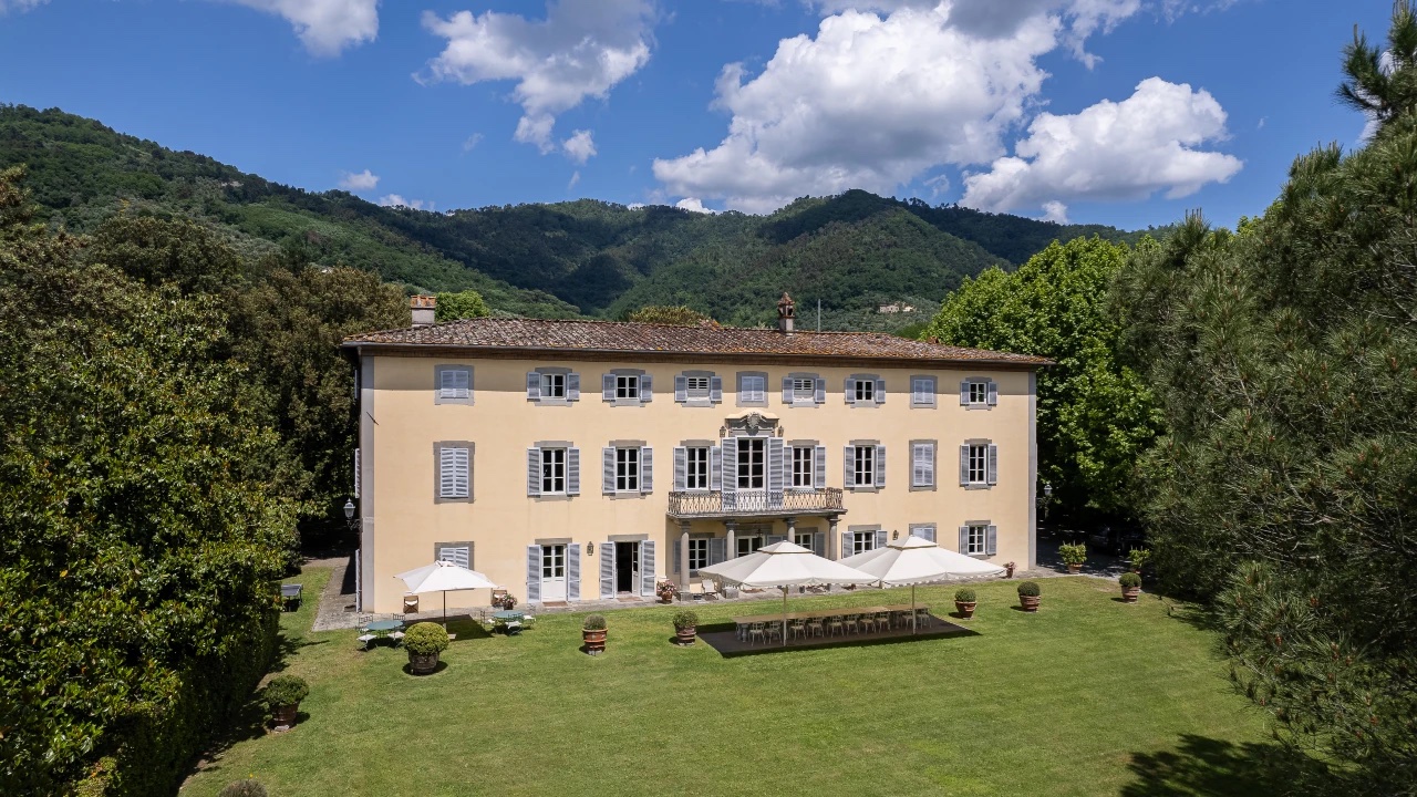 A large, light yellow villa reminiscent of Tuscany, with numerous windows and a tiled roof, stands on a manicured lawn surrounded by trees and hills under a blue sky. White umbrellas and outdoor seating are set up in front.