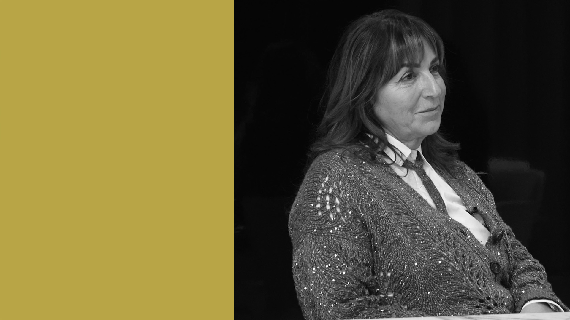 Una donna con capelli di media lunghezza che indossa un maglione strutturato e abbottonato è seduta a un tavolo e guarda di lato. Lo sfondo è nero, con un grande rettangolo verticale giallo sul lato sinistro dell'immagine.