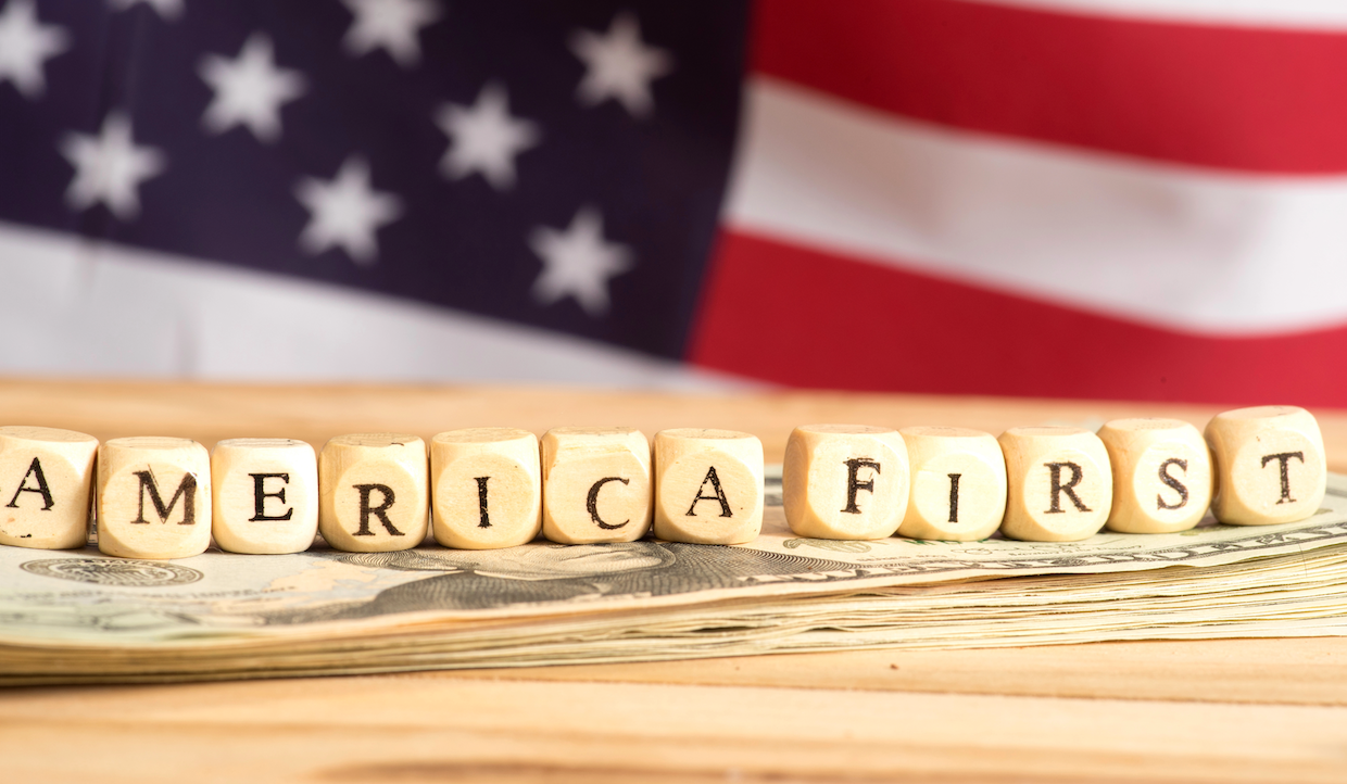 Blocchi di lettere in legno con la scritta "AMERICA FIRST" sono disposti sopra banconote da un dollaro americano, con una bandiera americana sfocata sullo sfondo.