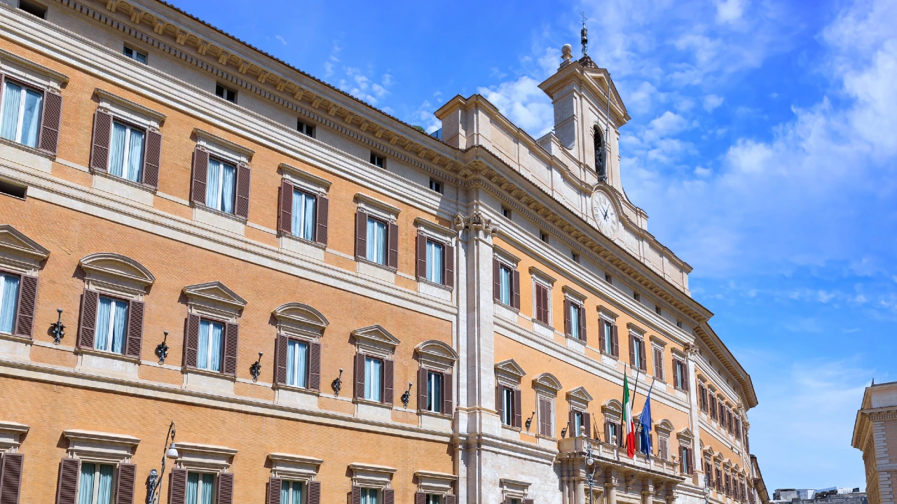 Un edificio governativo storico con mattoni beige, finestre ad arco e una torre dell'orologio centrale si erge sotto un cielo blu con nuvole sparse. Bandiere italiane e insegne relative al ddl conti correnti sono visibili vicino all'ingresso.