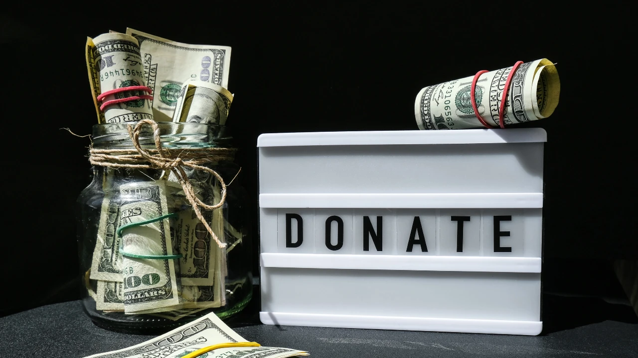 Un barattolo di vetro pieno di banconote arrotolate si trova accanto a una legittima insegna luminosa con la scritta "donate". Altri contanti sono arrotolati con elastici e banconote sciolte sono sparse sul tavolo. Il collegamento va al tema della donazione e della legittima, per cercare di capire quanto vale la quota ereditata