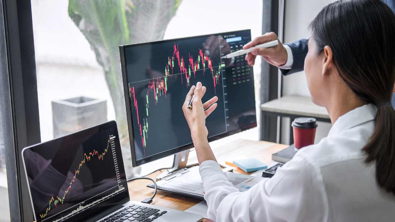 Due persone che analizzano grafici finanziari su un monitor di computer e un portatile, indicando lo schermo con penne in un ufficio con documenti e una tazza di caffè sulla scrivania.