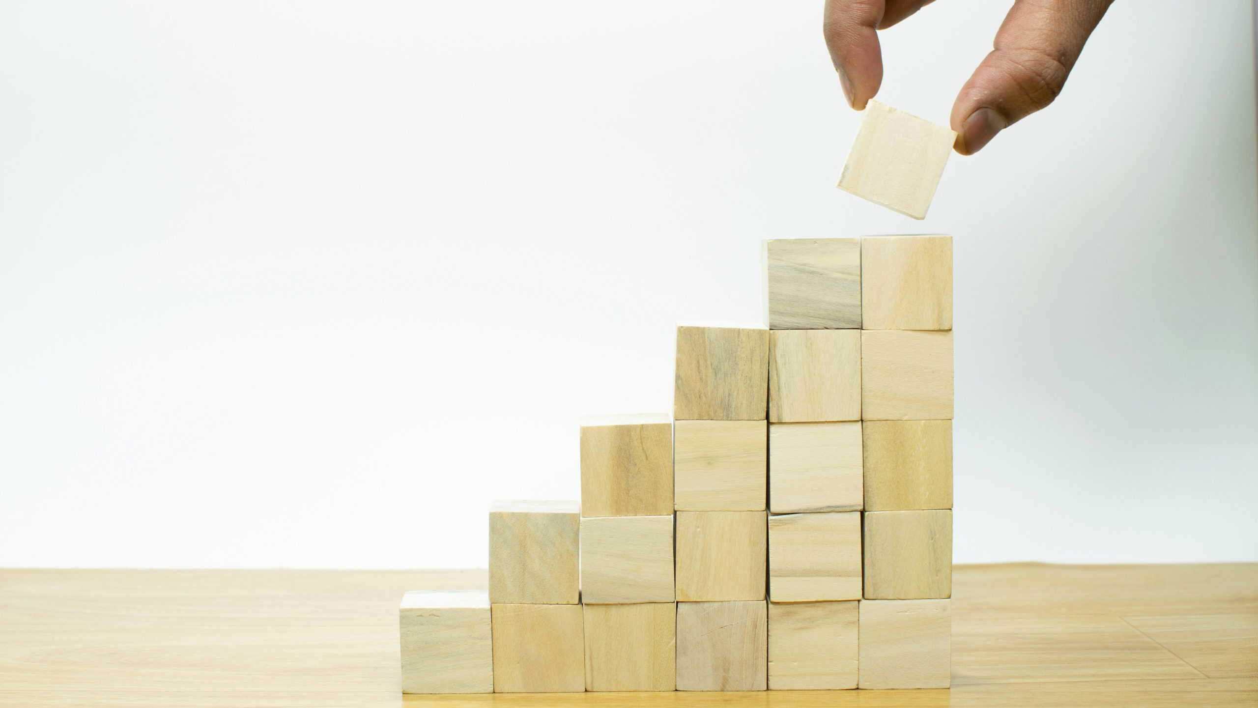 Una mano posiziona un blocco di legno in cima a una pila di blocchi di legno simili a scale, che simboleggiano la crescita, il progresso o la costruzione passo dopo passo, su una superficie di legno con uno sfondo bianco.