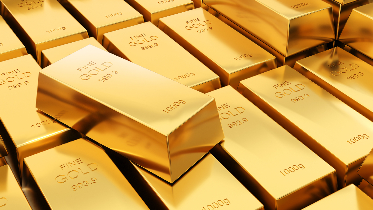 Numerosi lingotti d'oro lucenti sono impilati insieme, ciascuno etichettato come "ORO FINE 999,9" e "1000g". Un lingotto d'oro è posizionato in modo evidente sopra gli altri.
