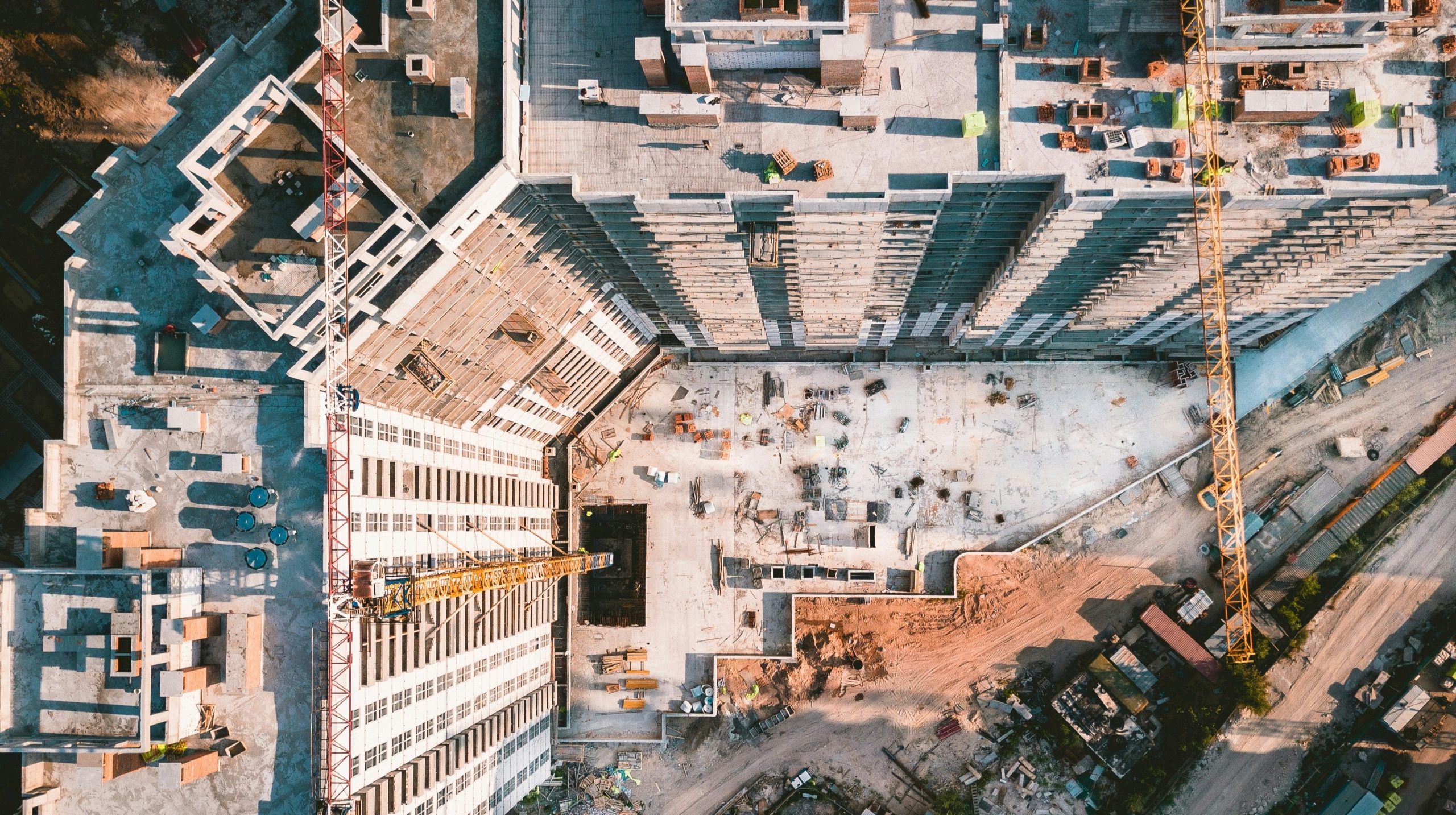Veduta aerea di un cantiere che mostra un grattacielo parzialmente costruito, gru, materiali da costruzione e fondamenta in calcestruzzo a vista circondate da terra e macchinari.