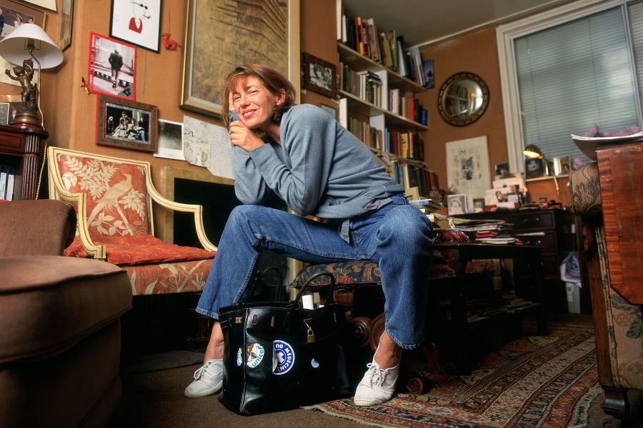 Una donna in maglione blu e jeans siede sorridente su una sedia in una stanza accogliente e disordinata, piena di libri, opere d'arte e oggetti personali, con una grande borsa nera ai suoi piedi che ricorda l'iconica borsa Jane Birkin.