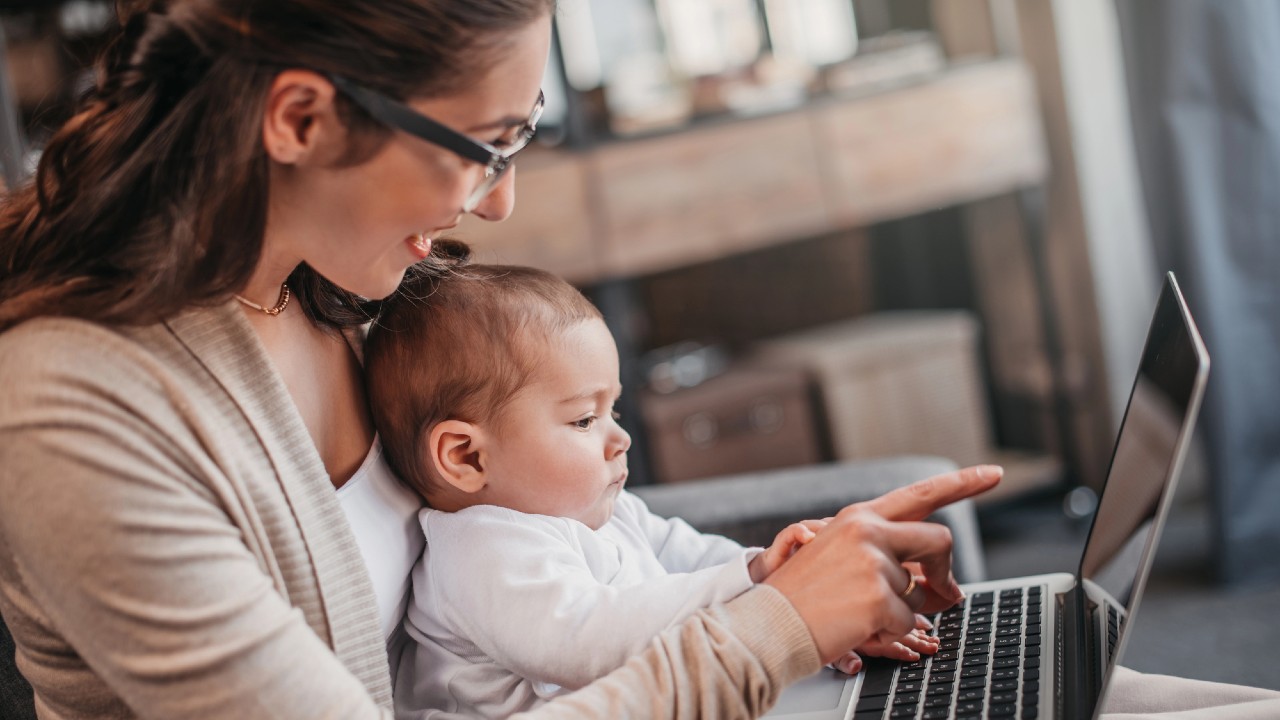 Una donna con occhiali e cardigan beige tiene in grembo un bambino mentre utilizza un computer portatile, conciliando lavoro e maternità obbligatoria. Il bambino guarda lo schermo e si avvicina alla tastiera mentre la donna sorride e indica il computer portatile.