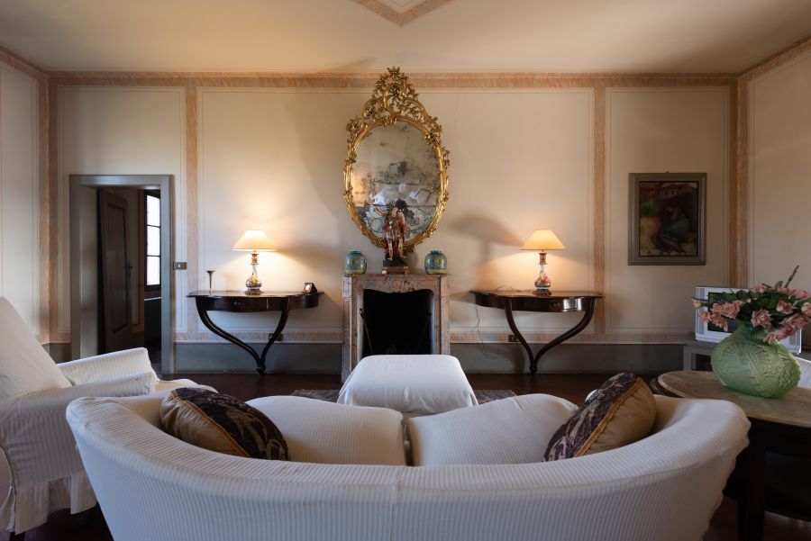 Il soggiorno, arredato con gusto nello stile di una villa di Lina Wertmüller, presenta un divano bianco e poltrone di fronte al camino. Sopra c'è uno specchio ornato da una cornice dorata, con una luce calda proveniente da lampade e un vaso di fiori su un tavolino.