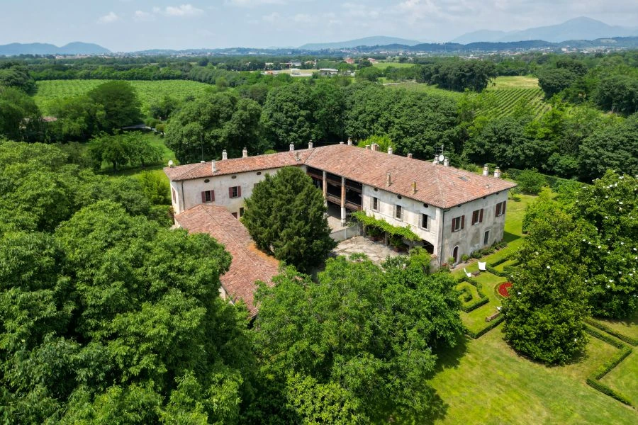 Veduta aerea della villa di Lina Wertmüller, una grande tenuta storica con il tetto di tegole rosse, immersa nel verde, tra giardini, alberi e montagne lontane, sotto un cielo limpido.