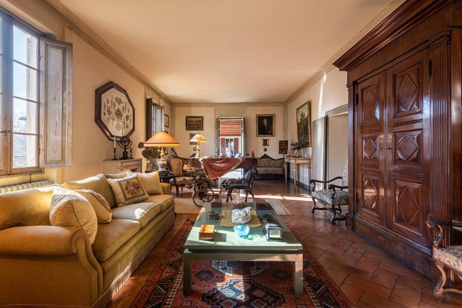 Ampio soggiorno illuminato dal sole con divano giallo, tavolino in legno, grande armadio, pianoforte e decorazioni antiche. Il pavimento in cotto e il tappeto persiano evocano il fascino di una villa di Lina Wertmüller, creando un'atmosfera calda e accogliente.