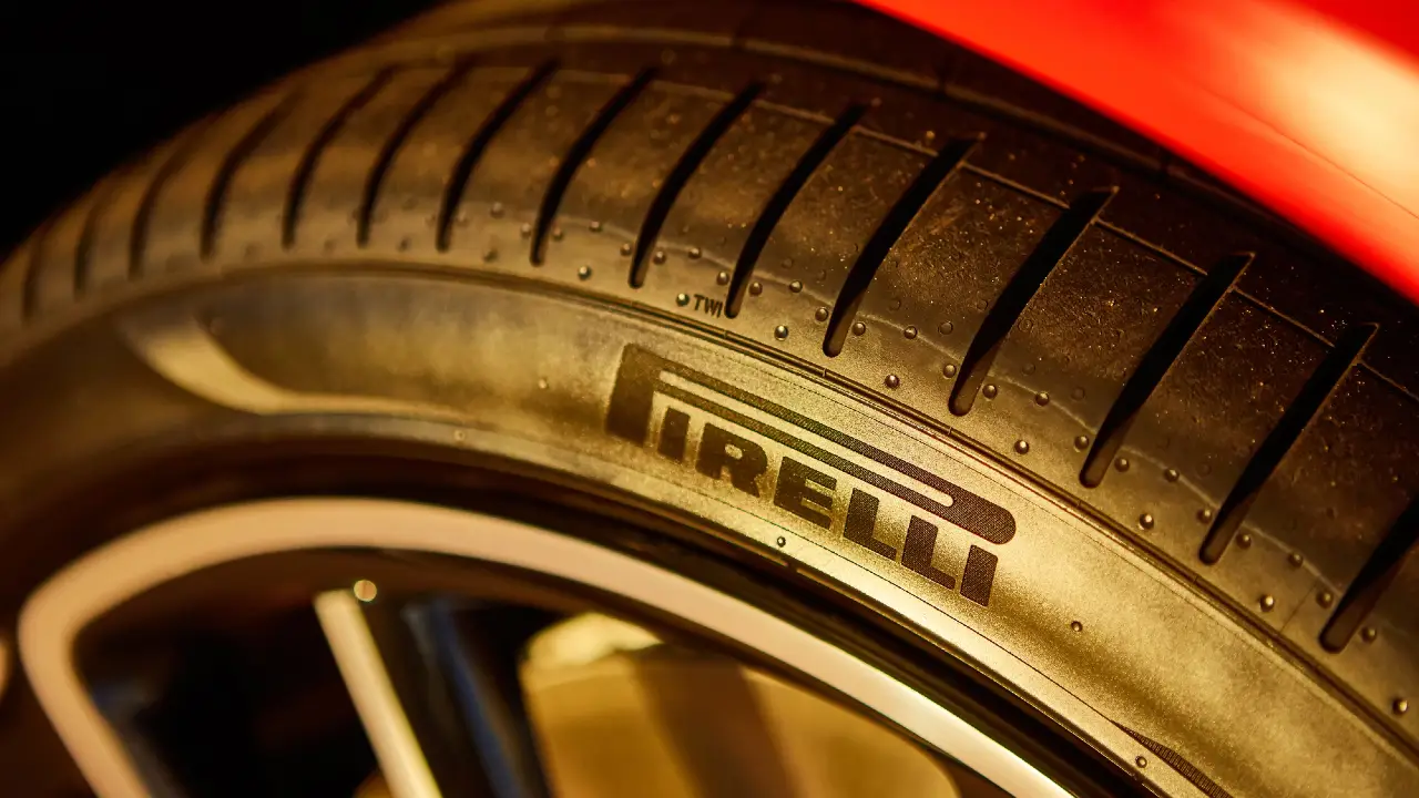 Primo piano di un pneumatico Pirelli nero per auto che mostra il logo del marchio sul fianco, con parte della carrozzeria di un veicolo rosso visibile sullo sfondo.