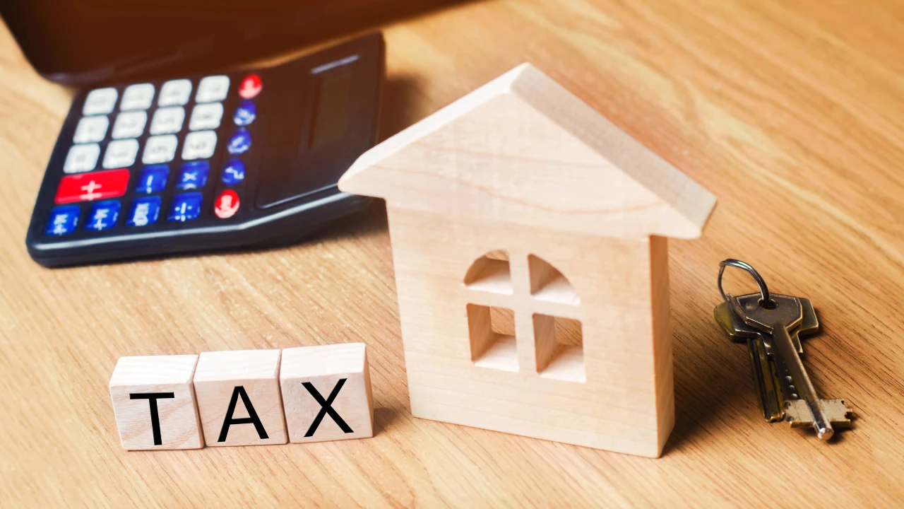 Un modello di immobile in legno, una calcolatrice, un mazzo di chiavi e blocchi di legno con la scritta "Tax" sono disposti su un tavolo di legno, a simboleggiare l'imposta sulla proprietà o la tassazione immobiliare sulle plusvalenze.