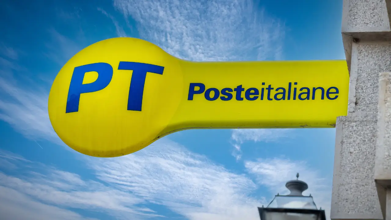 Un'insegna gialla con le lettere blu "PT" e "Poste italiane", che indica un ufficio postale italiano, contro un cielo blu con nuvole sparse.