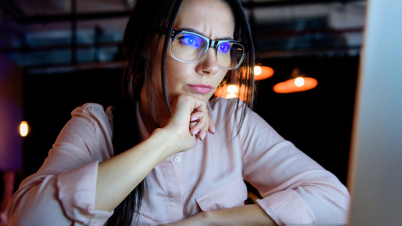 Una donna con gli occhiali siede di notte davanti allo schermo di un computer, appoggiando il mento sulla mano e guardando con attenzione. Sullo sfondo brillano luci calde.