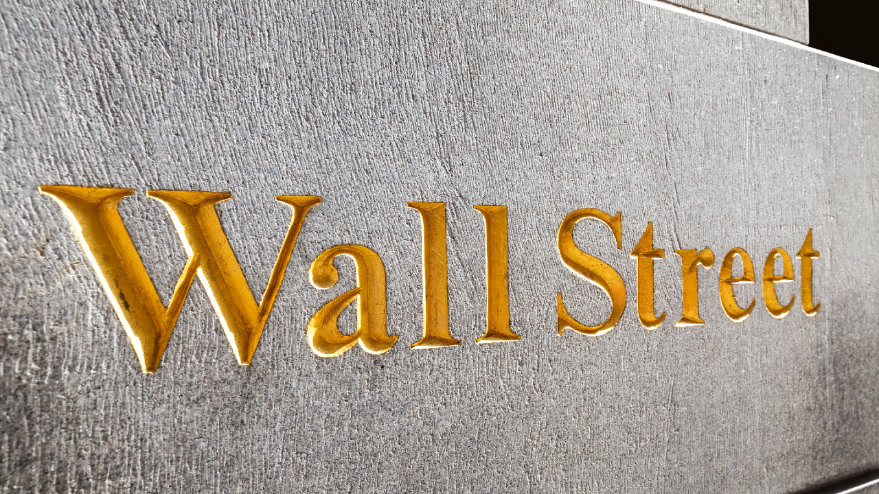 L'immagine mostra le parole "Wall Street" incise in caratteri dorati su un muro di pietra grigio.