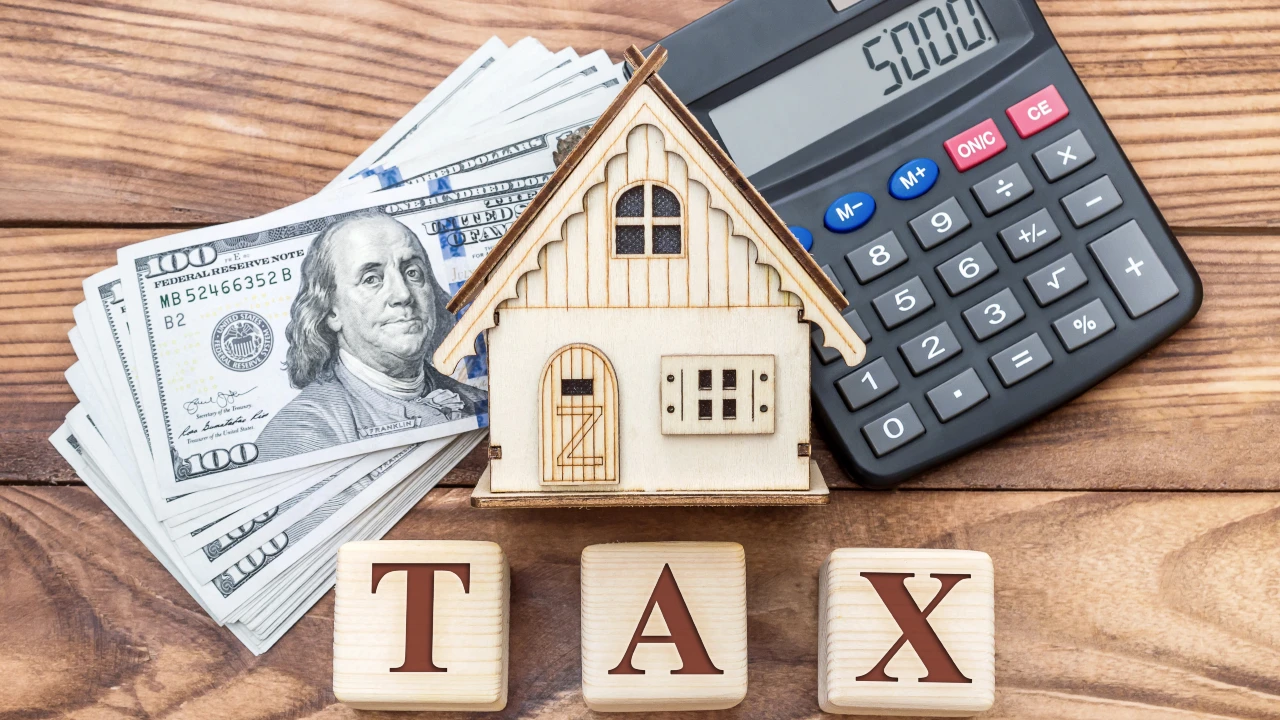 Su una superficie di legno sono disposti un modello di casa in legno, una pila di banconote da cento dollari, una calcolatrice che indica "5000" e blocchi di lettere in legno che scrivono "Tax". Il riferimento va alla tassazione della plusvalenza da trust esteri su immobili in Italia.