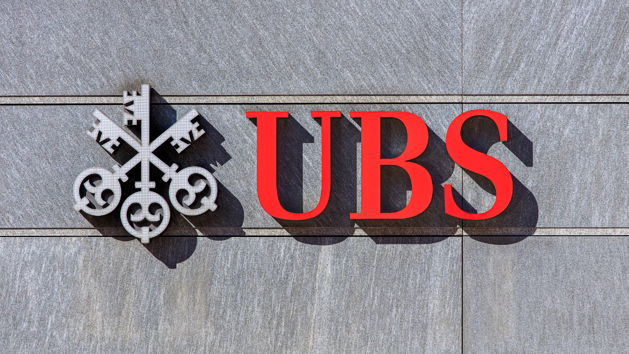 Il logo e il nome di UBS, in grassetto rosso, appaiono su una parete di pietra grigia, con l'emblema di una chiave sul lato sinistro.