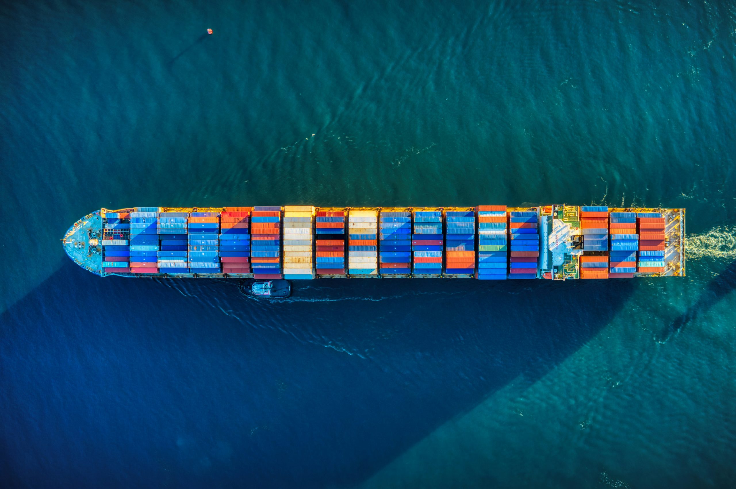 Vista aerea di una grande nave da carico con container colorati che naviga nell'acqua blu profonda, creando una scia dietro di sé.