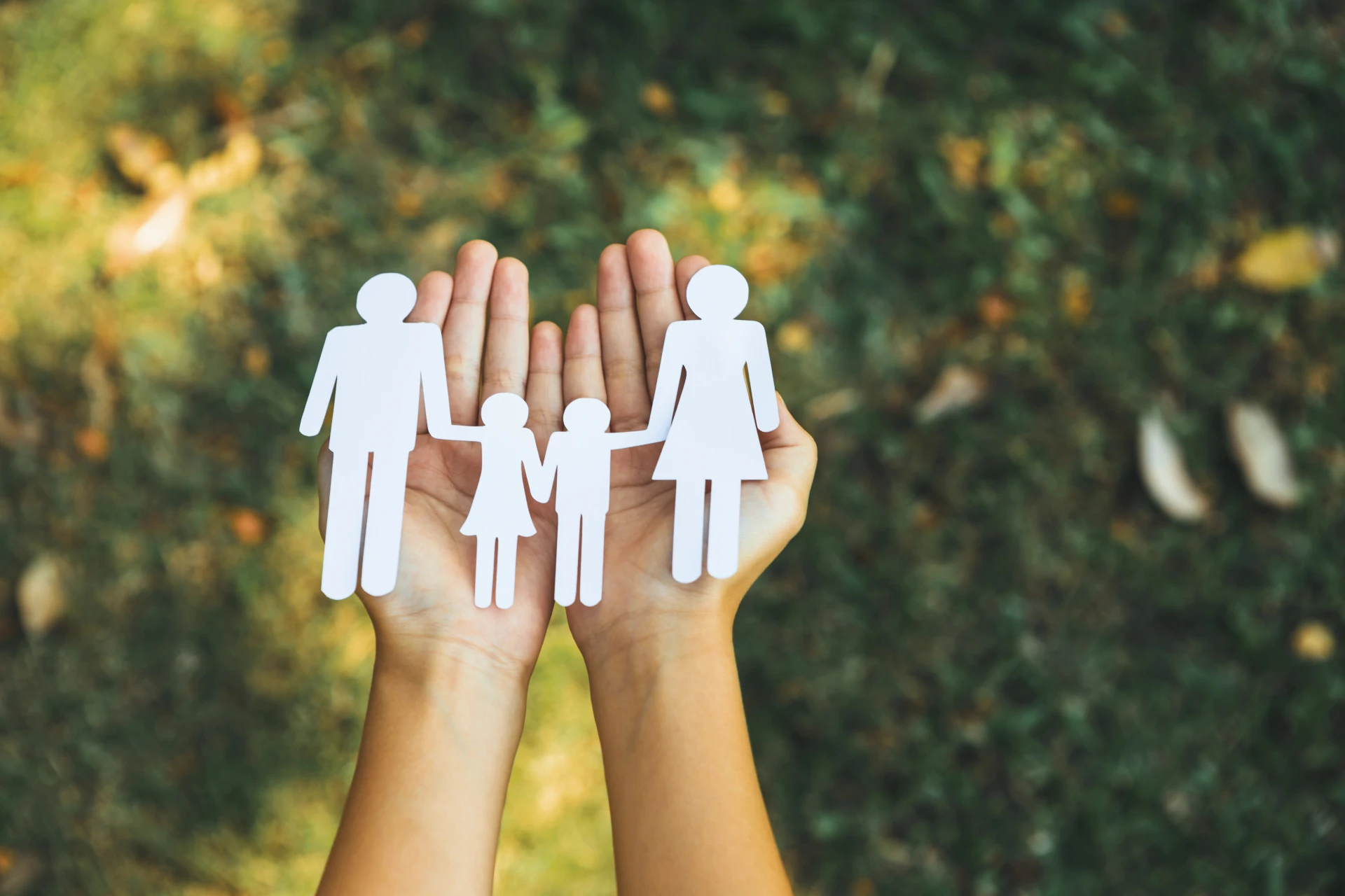 Un paio di mani reggono i ritagli di carta di una famiglia - due adulti e due bambini - su uno sfondo erboso sfocato.