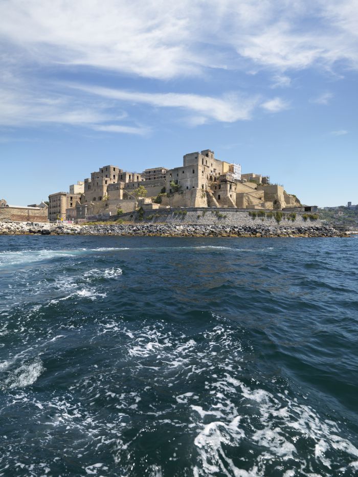Una fortezza in pietra con diversi edifici si trova su una costa rocciosa sotto un cielo parzialmente nuvoloso, vista dall'acqua, con le onde in primo piano: un suggestivo panorama di Pozzuoli.