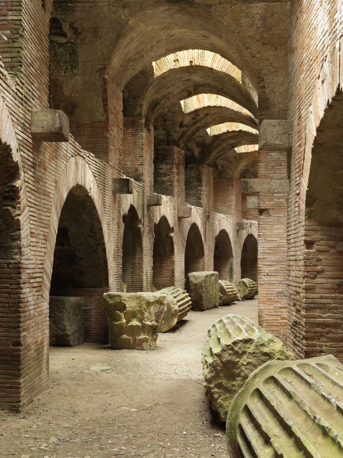 Gli antichi archi in mattoni e i soffitti a volta di un rudere romano, con grandi colonne di pietra spezzate sparse sul terreno, sono illuminati dalla luce naturale dall'alto e offrono un panorama suggestivo dell'affascinante storia di Pozzuoli.