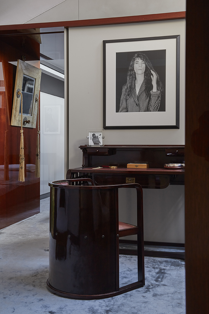 Un elegante ufficio domestico evoca la raffinata eleganza della Casa Museo Molinario Colombari, con una scrivania in legno scuro lucido e una sedia curva abbinata su un tappeto grigio chiaro. Un ritratto in bianco e nero e caldi accenti in legno completano questo spazio moderno.
