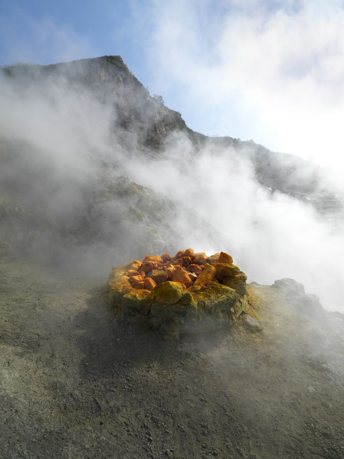 Un mucchio di rocce sulfuree di colore giallo e arancione, circondate da vapore, si trova su un terreno roccioso vicino a una montagna nebbiosa, catturando la suggestiva bellezza naturale di Panorama Pozzuoli.