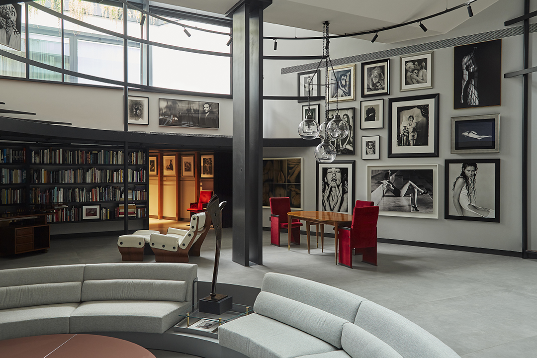 Un soggiorno moderno che ricorda la casa museo molinario colombari è caratterizzato da divani curvi, un tavolino rotondo e sedie di velluto rosso attorno a un tavolo di legno. Ritratti in bianco e nero adornano le pareti; grandi finestre e librerie completano l'ambiente.