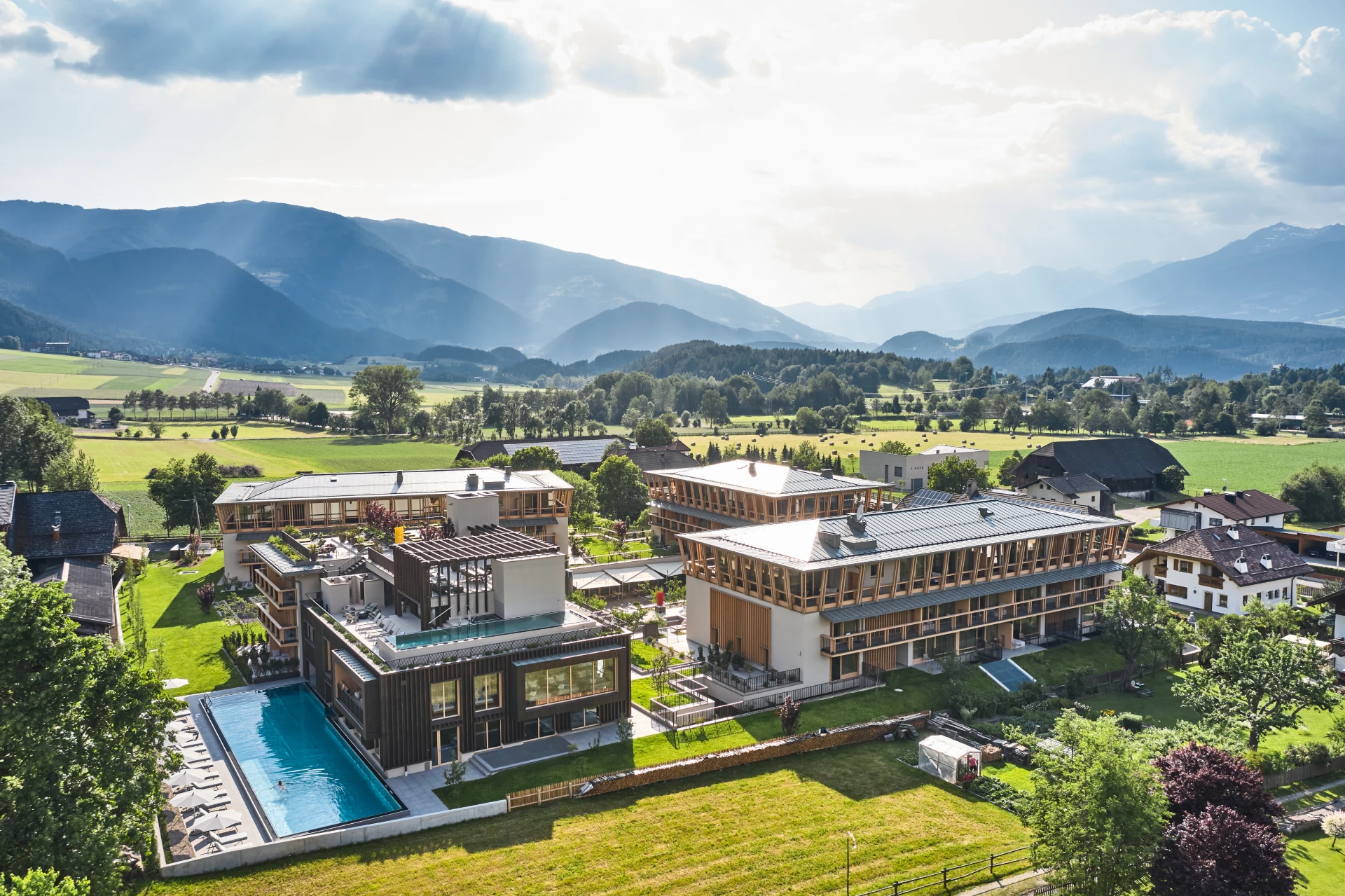 Un moderno complesso alberghiero con piscina all'aperto immerso in una valle verde, circondato da alberi, campi e montagne sotto un cielo parzialmente nuvoloso con raggi di sole che si fanno strada.