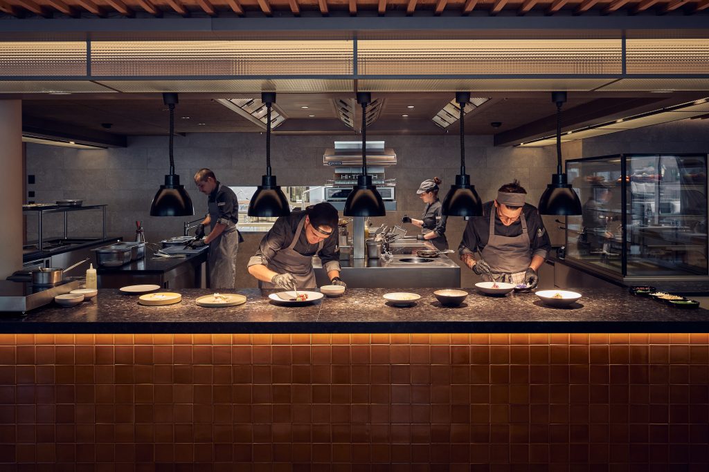 Gli chef del Falkensteiner Hotel Kronplatz lavorano in una moderna cucina aperta, impiattando i piatti sotto lampade nere. Concentrati sui banchi in acciaio inox, preparano i pasti con le eleganti attrezzature da cucina visibili sullo sfondo.