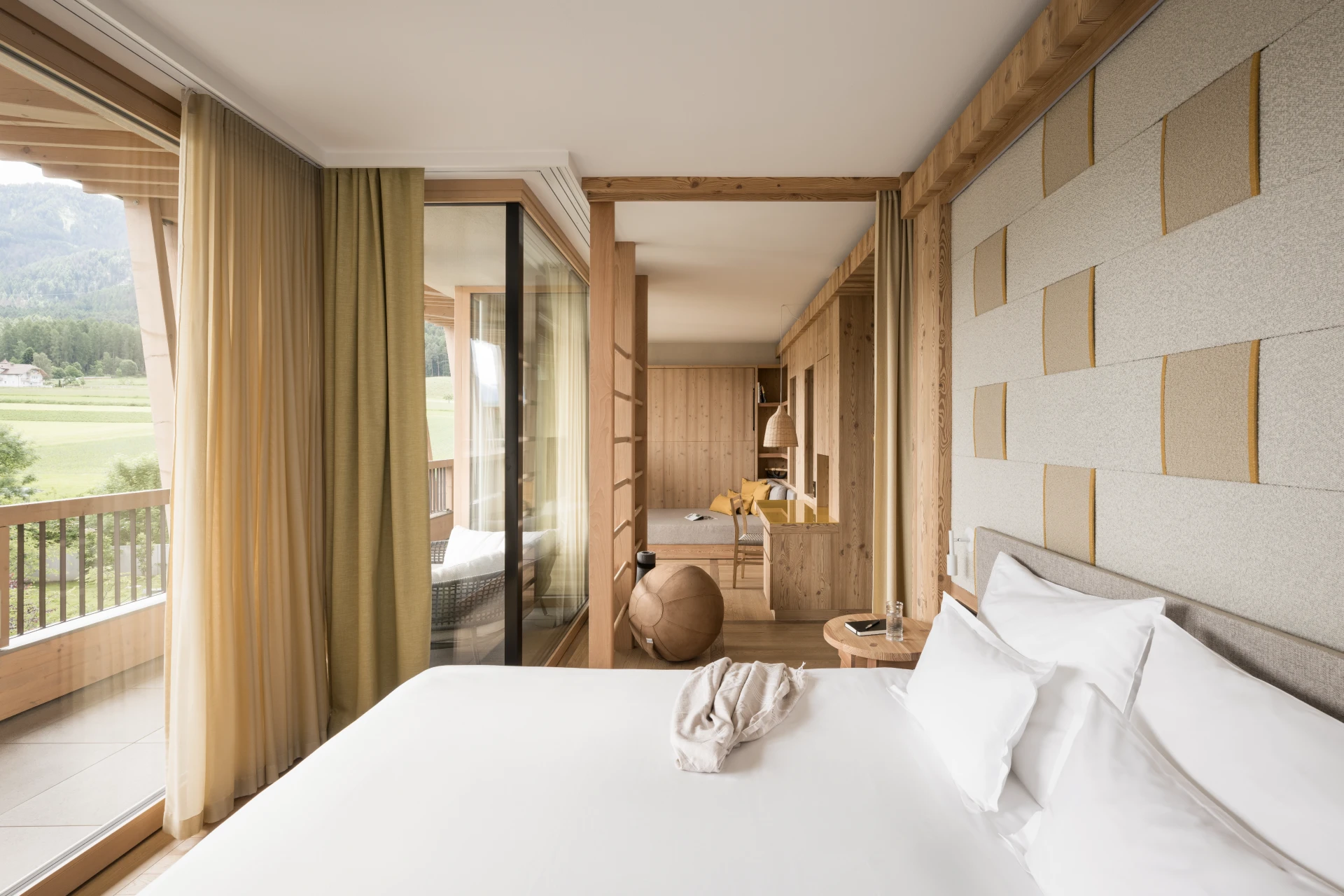 Una moderna camera del Falkensteiner Hotel Kronplatz con un grande letto, biancheria bianca, arredi in legno e una parete di vetro che conduce a un balcone con vista sulle montagne. L'ambiente è luminoso grazie alla luce naturale e ai toni caldi e neutri.