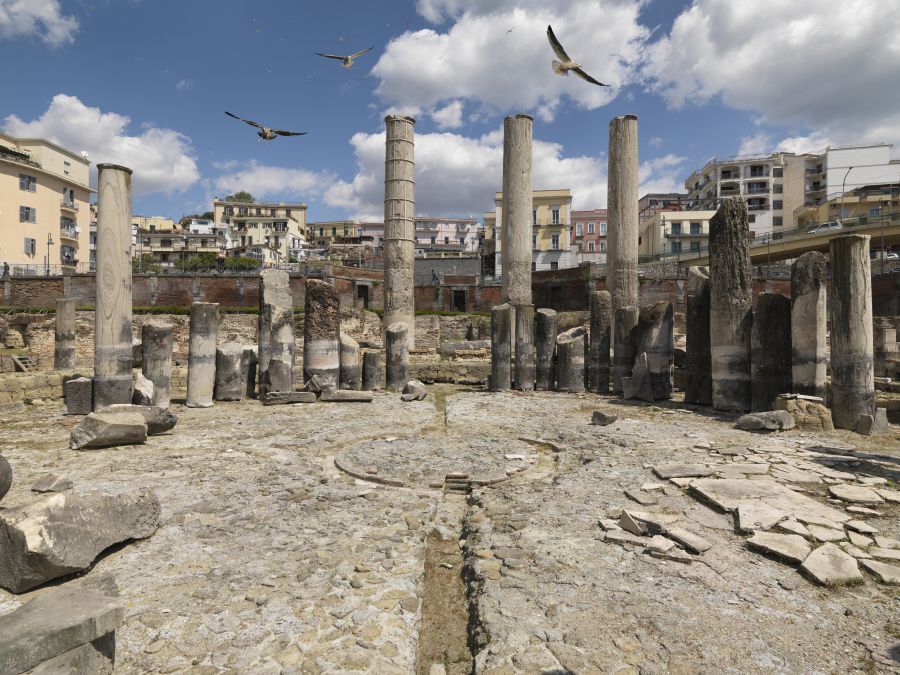 Antiche colonne di pietra e rovine sotto un cielo parzialmente nuvoloso, con gabbiani che volano sopra la testa ed edifici moderni sullo sfondo, creano un Panorama Pozzuoli mozzafiato.