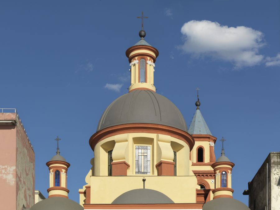 Un panorama di Pozzuoli con una chiesa a cupola gialla e rossa con croci in cima a torri rotonde e finestre ad arco, in un cielo azzurro e limpido con un'unica nuvola bianca.