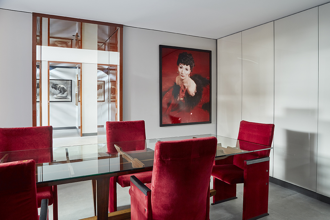 Una sala da pranzo moderna con un tavolo di vetro circondato da sedie di velluto rosso, pareti bianche e un grande ritratto di donna in rosso e nero esposto alla parete, che evoca l'ambiente raffinato della Casa Museo Molinario Colombari. Una porta rivela altre opere d'arte nelle vicinanze.