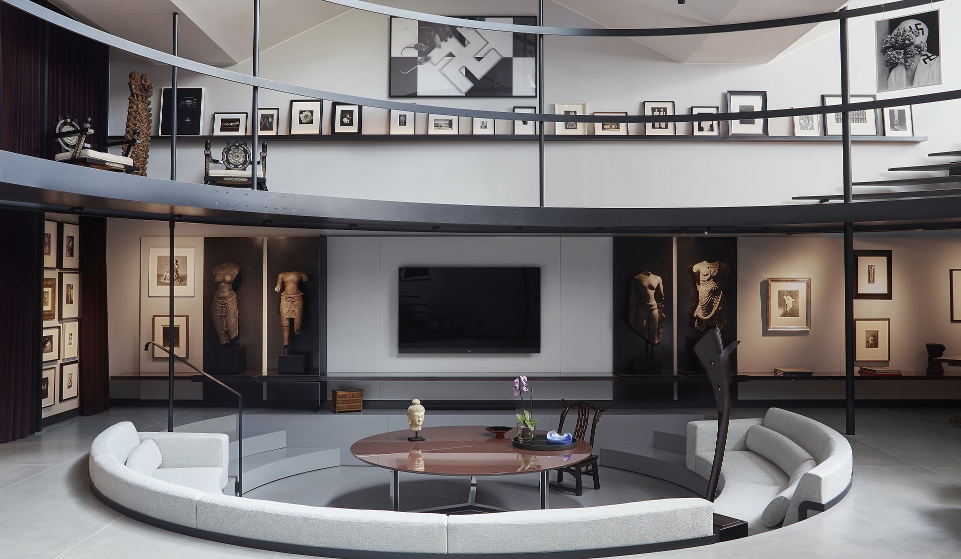 Soggiorno moderno con zona salotto circolare incassata, tavolino rotondo, TV a parete e soppalco rivestito di opere d'arte e libri, che richiama l'atmosfera da galleria della casa museo Molinario Colombari.