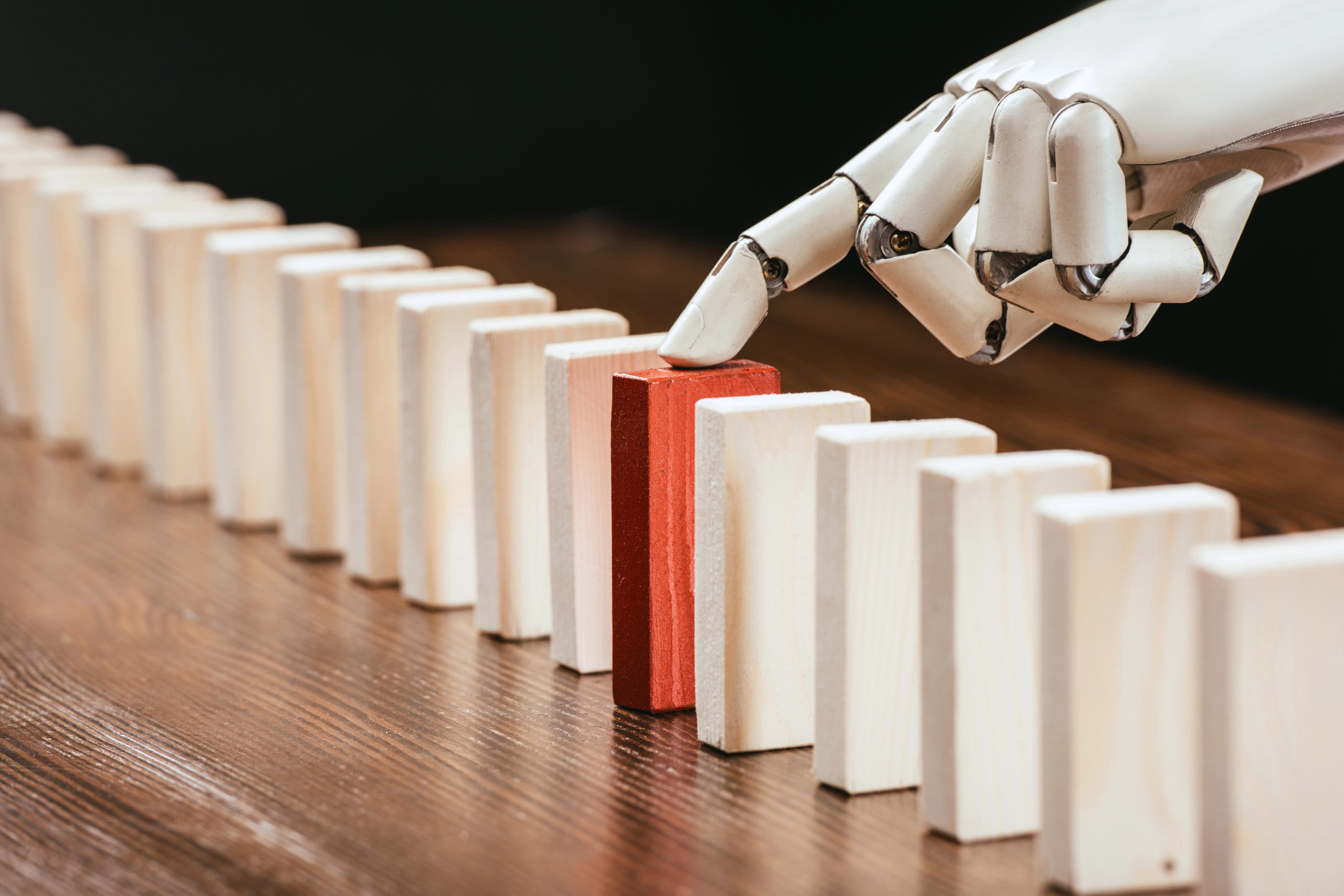 Una mano robotica ferma una fila di domino bianchi in caduta toccando un singolo domino rosso su un tavolo di legno, simboleggiando l'intervento o il controllo: una metafora appropriata per la precisione e la supervisione offerte dalla tecnologia dei family office.